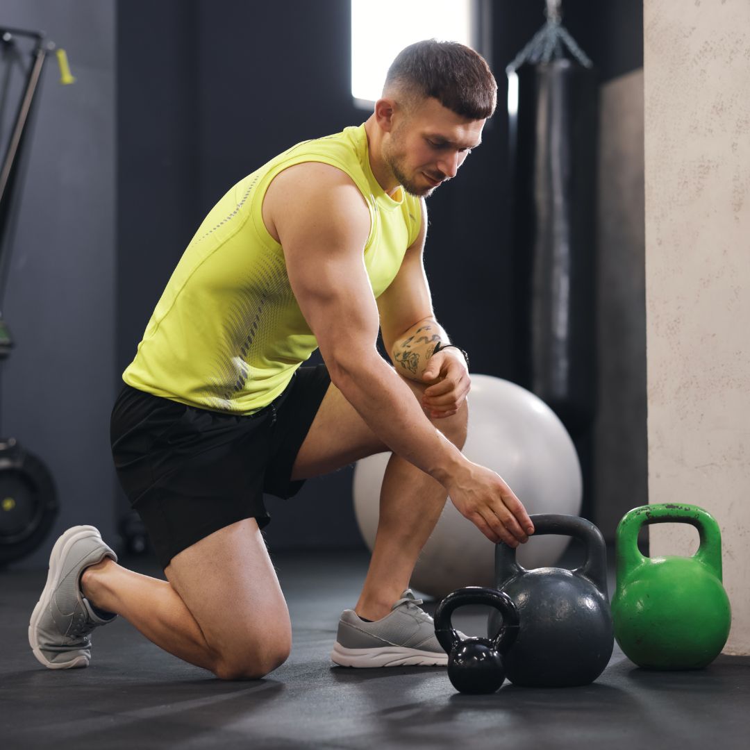 Muscular man in yellow tank top kneeling with kettlebells during intense gym workout