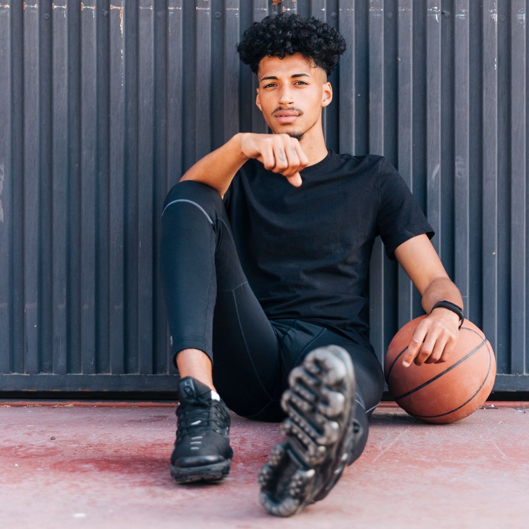 Confident young man sitting with basketball against urban wall in black athletic wear